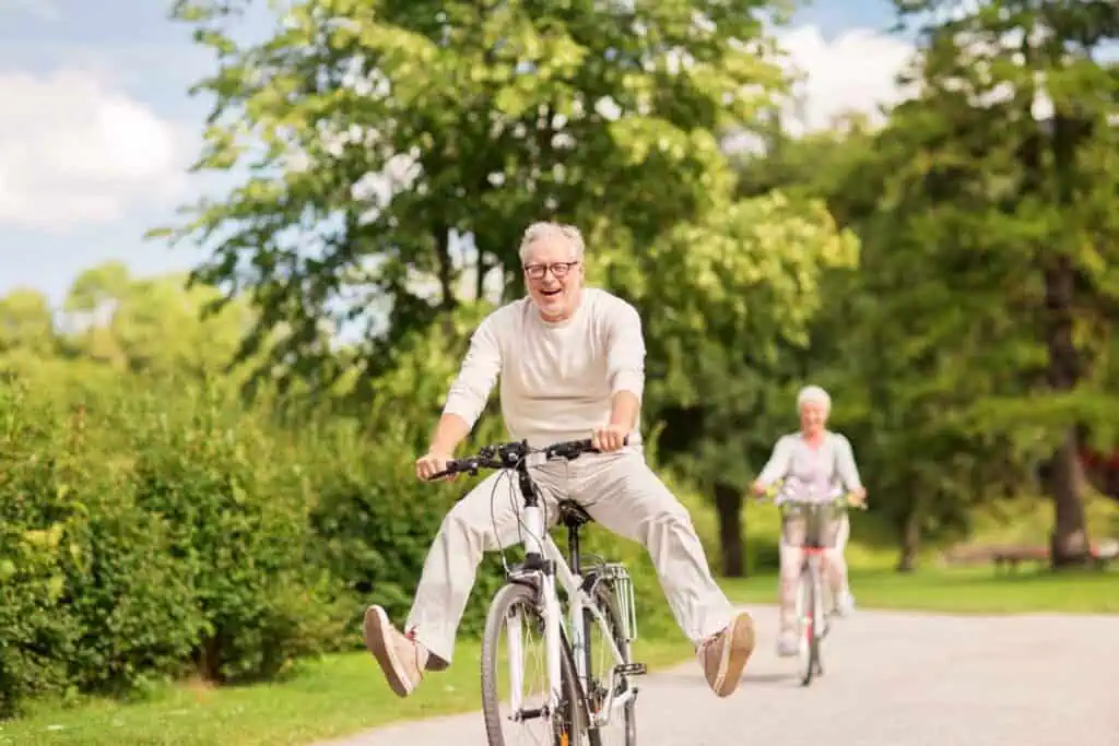 Ein fröhlicher älterer Mann fährt Fahrrad mit von den Pedalen gestreckten Beinen, während eine ältere Frau auf einem Fahrrad im Hintergrund fährt, alles in einem grünen Park.