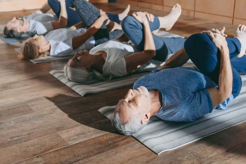Vier ältere Erwachsene liegen auf dem Rücken auf Yogamatten und ziehen ihre Knie zur Brust, um sich zu dehnen.