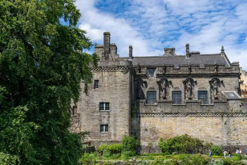 Stirling Castle in Schottland, eine große, alte Steinburg mit verschiedenen architektonischen Details wie Gesimsen und Verzierungen, flankiert von einem großen grünen Baum auf der linken Seite und einem blauen Himmel mit einigen Wolken.