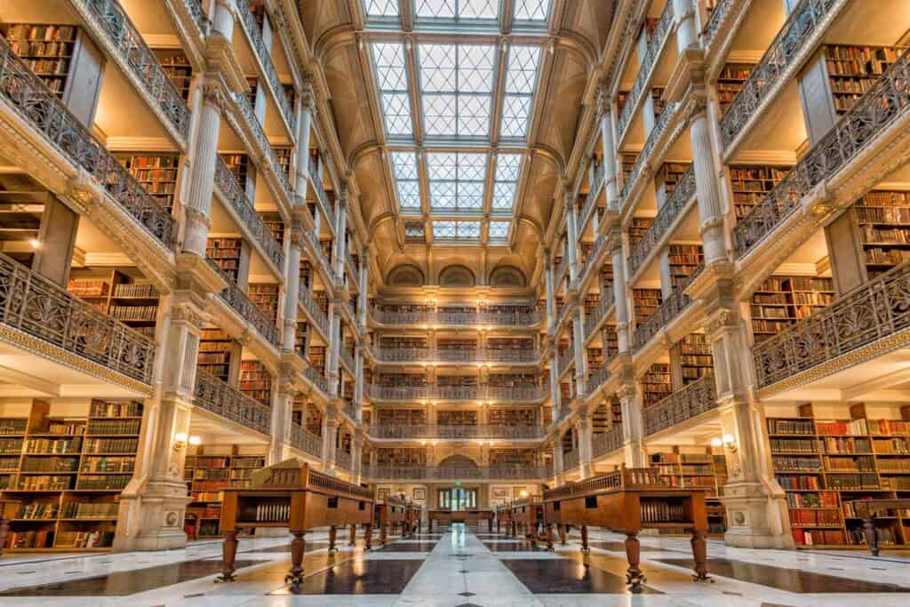 Das hohe Innere der George Peabody Library in Baltimore, Maryland, mit fünf Stockwerken voller Bücherregale, die von verzierten Gusseisenbalkonen gesäumt sind, die zu einem großen Oberlicht reichen. Auf dem Marmorboden stehen Holztische.