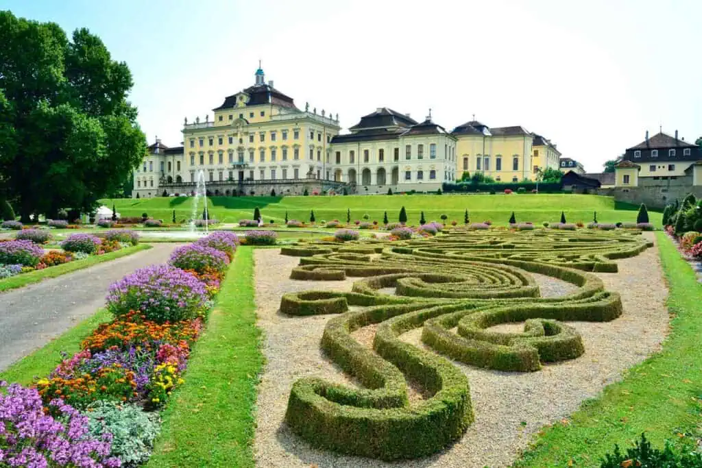 Die Schlossanlage Solitude in Stuttgart, Deutschland, mit einem großen, gelben Barockschloss auf einem Hügel, umgeben von gepflegten Gärten mit kunstvollen Formschnittbeeten, Blumenrabatten und einem Springbrunnen.