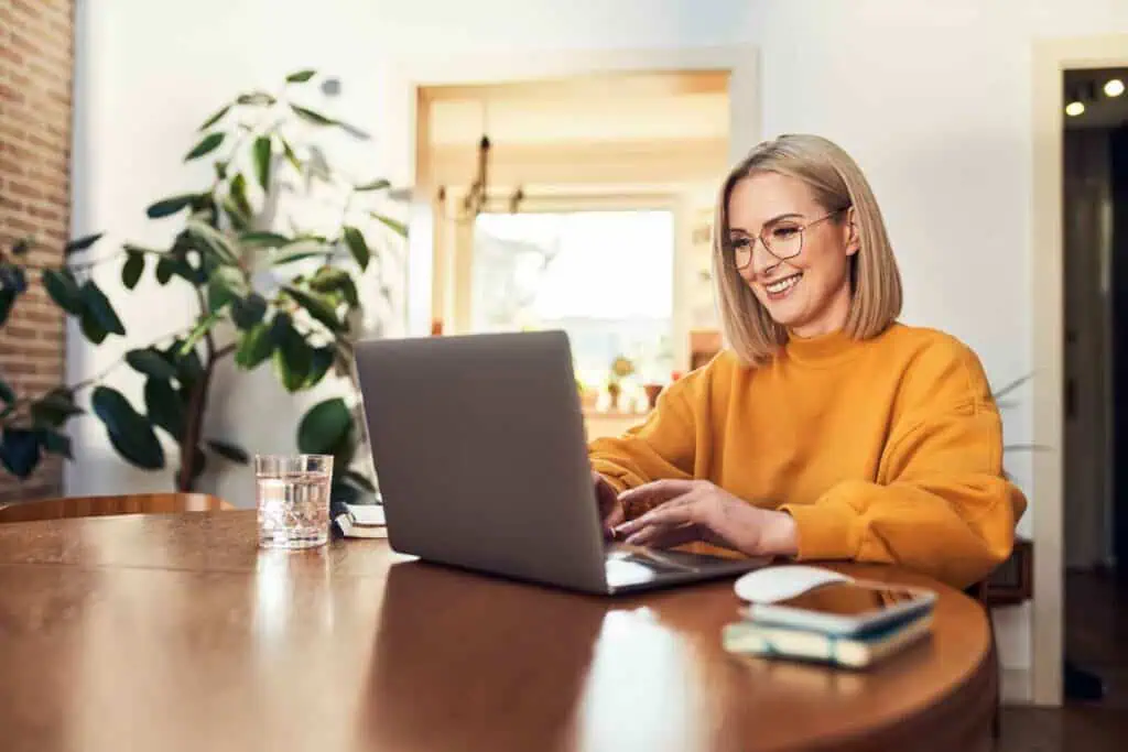 Eine lächelnde, blonde Frau mit Brille und einem gelben Pullover sitzt an einem Holztisch und tippt auf einem Laptop, neben dem ein Glas Wasser, eine Maus und ein Smartphone liegen.