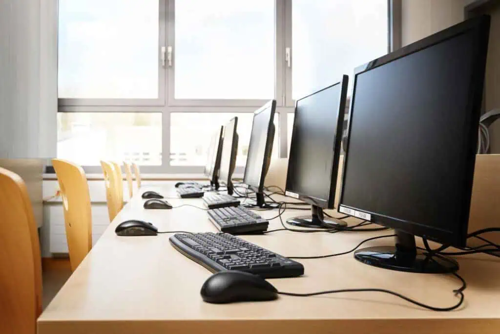 Fünf Computermonitore und -tastaturen, die in einer Reihe auf einem hellen Holztisch stehen, mit Stühlen, die davor positioniert sind. Im Hintergrund sind große Fenster zu sehen.