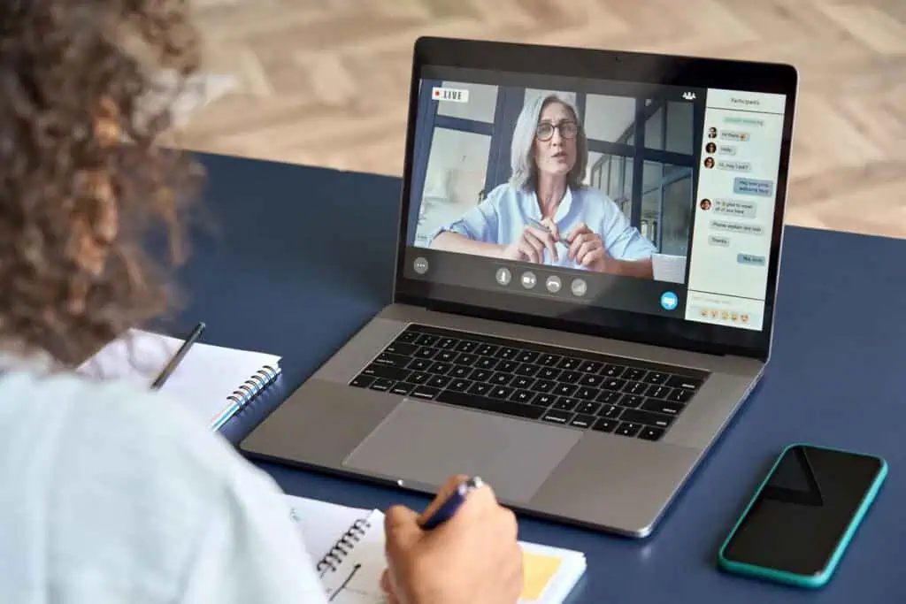 Eine Person sitzt an einem Schreibtisch und nimmt an einer Online-Videokonferenz oder einem Webinar auf einem Laptop teil, während sie Notizen in einem Notizbuch macht. Auf dem Bildschirm ist eine ältere Frau zu sehen, die spricht, und eine Chat-Leiste mit Teilnehmern und Nachrichten.