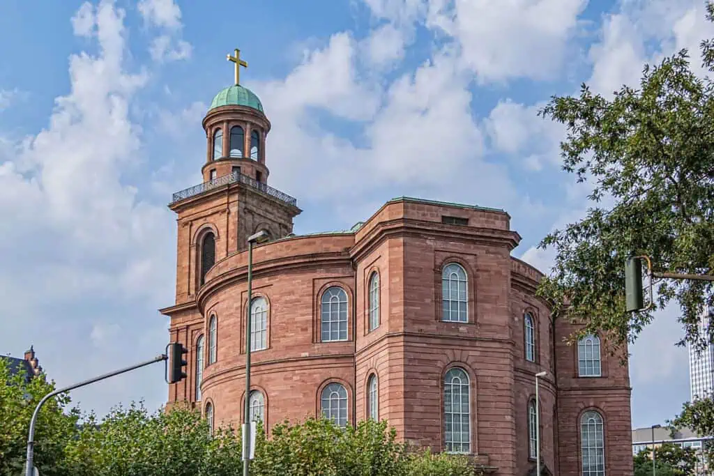 Die Paulskirche in Frankfurt am Main, ein rotes Sandsteingebäude mit einem runden Hauptteil und einem Turm mit grüner Kuppel und goldenem Kreuz, unter einem blauen Himmel mit weißen Wolken.