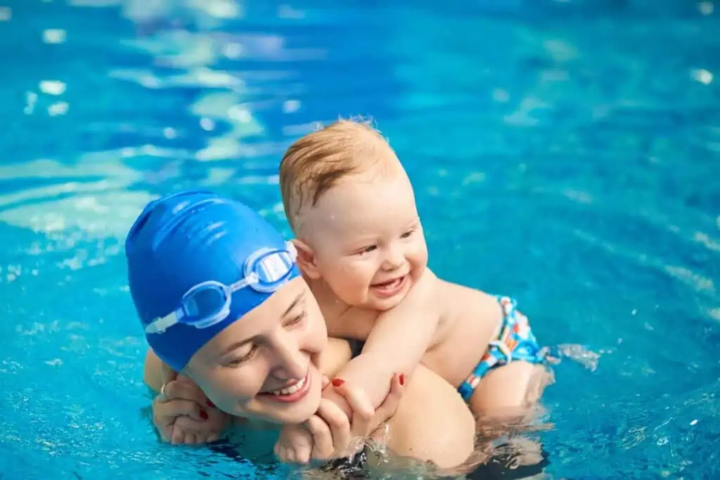 Eine lächelnde Frau mit blauer Badekappe und Schwimmbrille hält ein fröhliches Baby im Schwimmbecken.