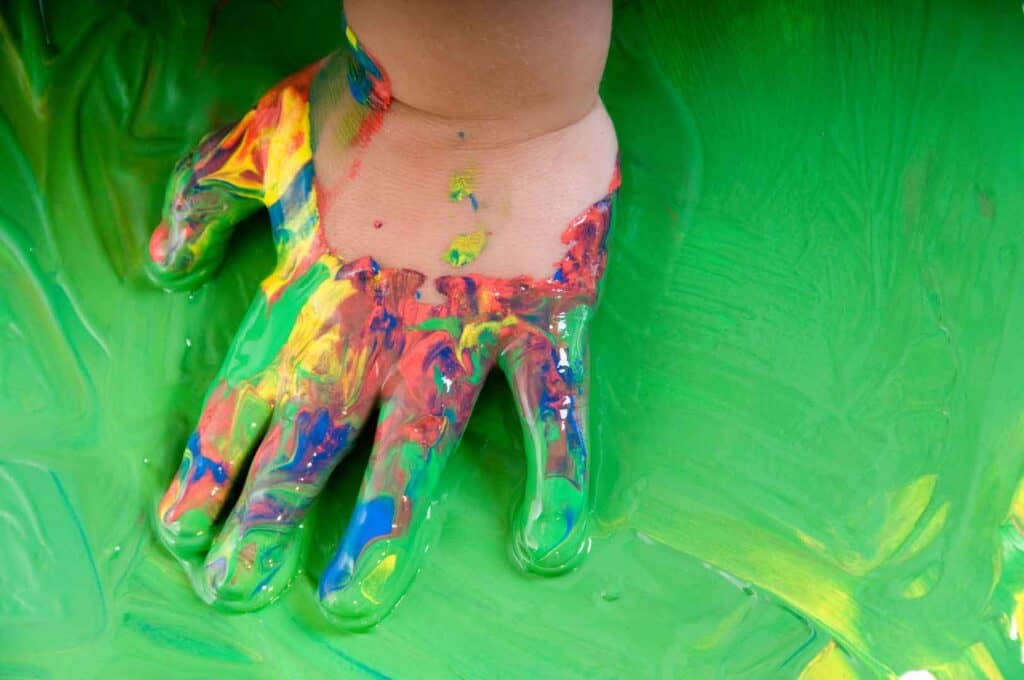 Nahaufnahme einer kleinen Kinderhand, die vollständig mit leuchtenden Farben wie Rot, Gelb, Blau und Grün bedeckt ist, und die in einem Schwall von grüner Farbe liegt.
