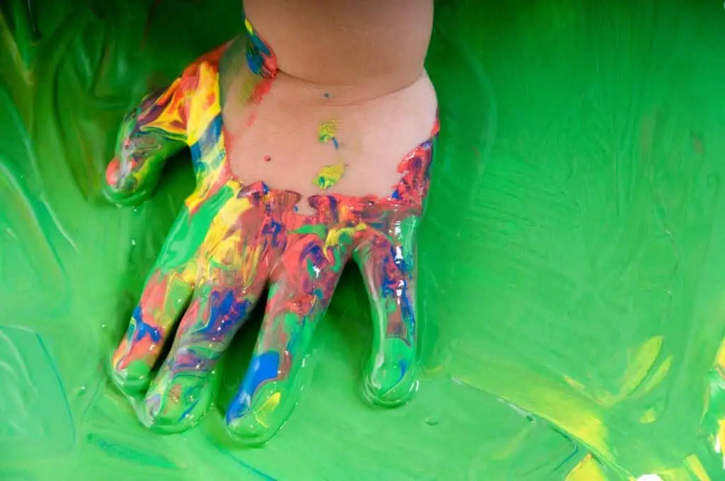 Nahaufnahme einer kleinen Kinderhand, die vollständig mit leuchtenden Farben wie Rot, Gelb, Blau und Grün bedeckt ist, und die in einem Schwall von grüner Farbe liegt.