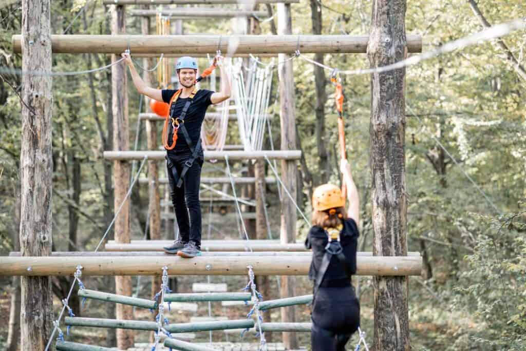 Ein Mann mit blauem Helm und Klettergurt steht lächelnd auf einer Holzplattform in einem Hochseilgarten, während eine weitere Person mit orangefarbenem Helm im Vordergrund zu sehen ist, umgeben von Bäumen.