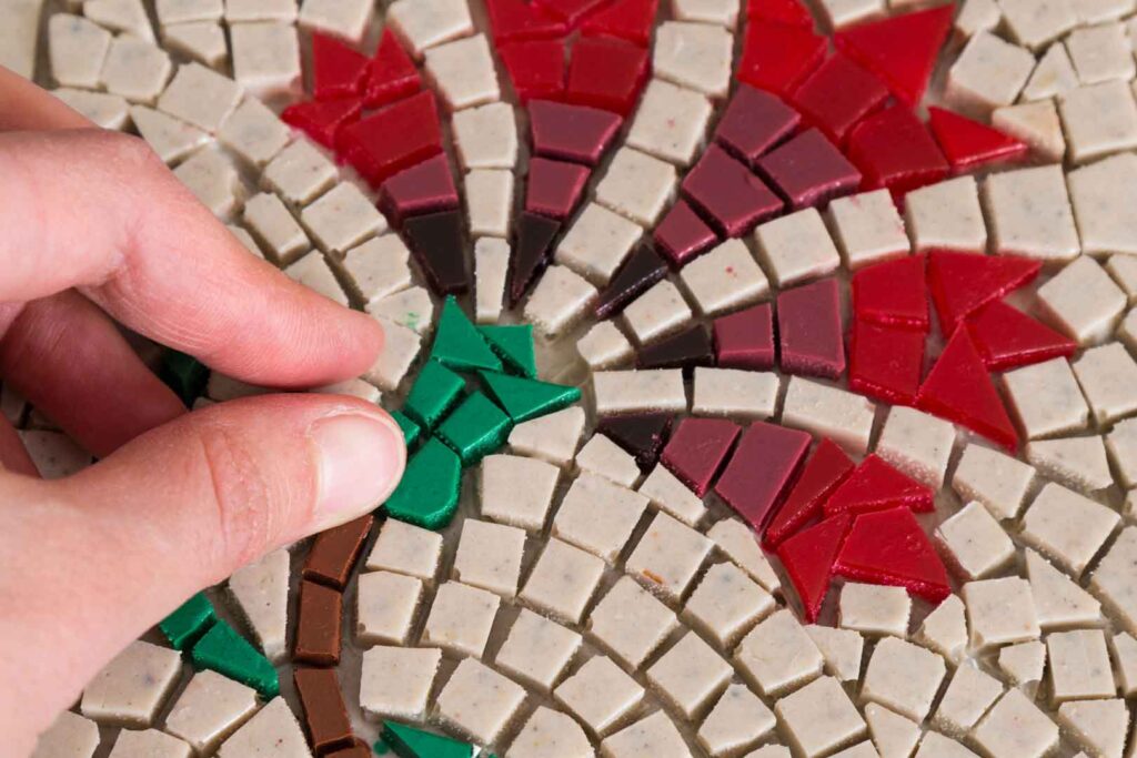 Eine Hand platziert ein grünes Mosaikstück auf einer Oberfläche, die mit vielen kleinen, quadratischen und dreieckigen Mosaikstücken in Creme, Rot und Dunkelrot belegt ist.