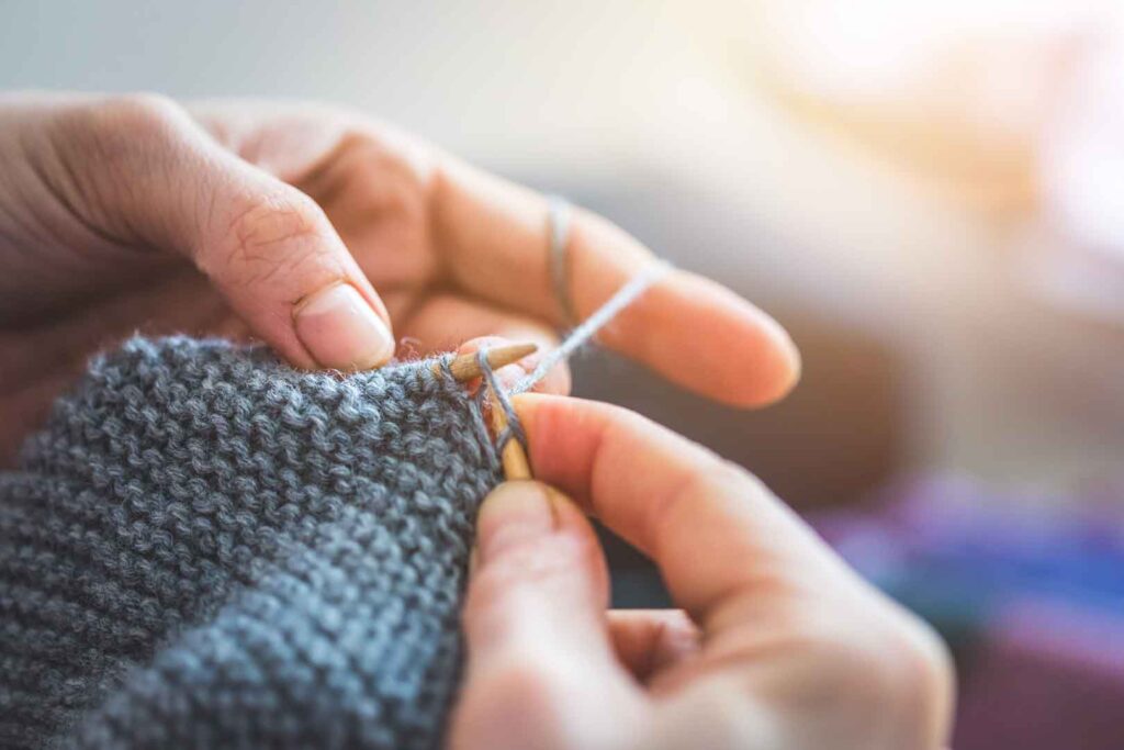 Hände stricken mit Holznadeln graue Wolle, wobei sich ein neues hellgraues Garn um den Finger wickelt und in die Maschen eingearbeitet wird, mit weichem Sonnenlicht von oben rechts.