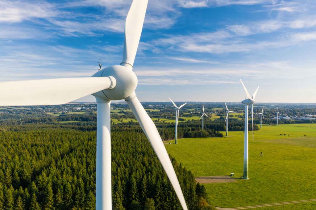 Ein Luftbild zeigt mehrere hohe Windturbinen, die sich über einem weitläufigen, grünen Feld und einem dichten Nadelwald erheben, unter einem blauen Himmel mit vereinzelten Wolken.