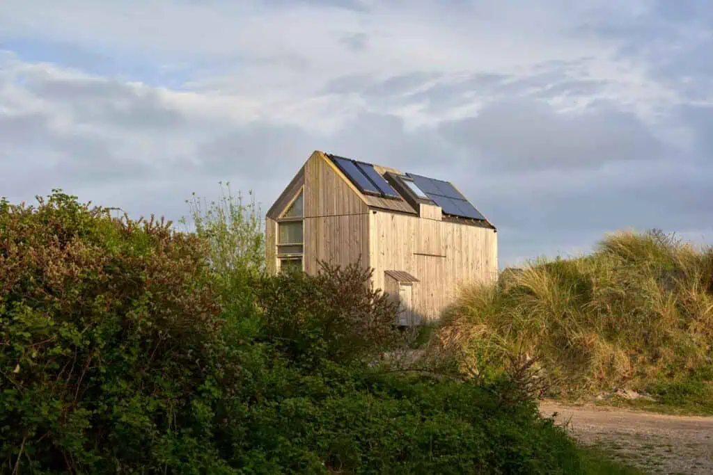 Ein kleines Holzhaus mit Solarpaneelen auf dem Dach, umgeben von grünen Büschen und Grashügeln unter einem bewölkten Himmel.
