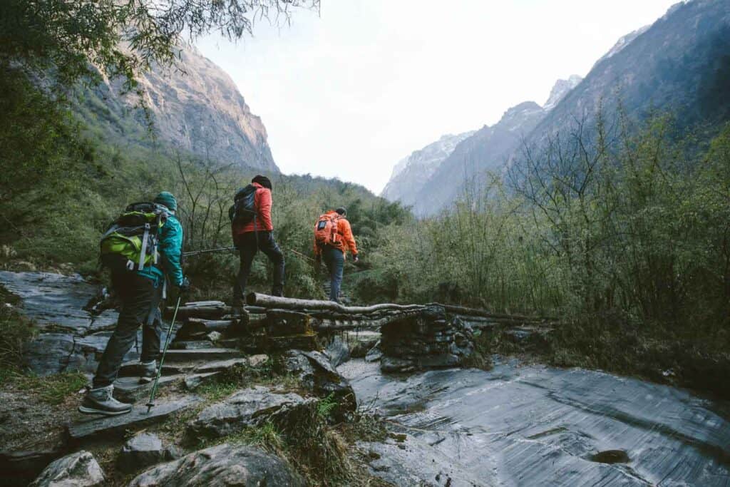 Drei Wanderer mit Rucksäcken überqueren eine kleine Holzbrücke in einem felsigen, bergigen Tal, umgeben von grüner Vegetation.