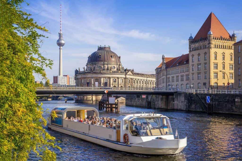 Ein Panoramablick auf die Berliner Stadtlandschaft, mit einem Touristenschiff auf einem Fluss im Vordergrund, dem Berliner Dom und dem Fernsehturm im Hintergrund unter einem klaren blauen Himmel.