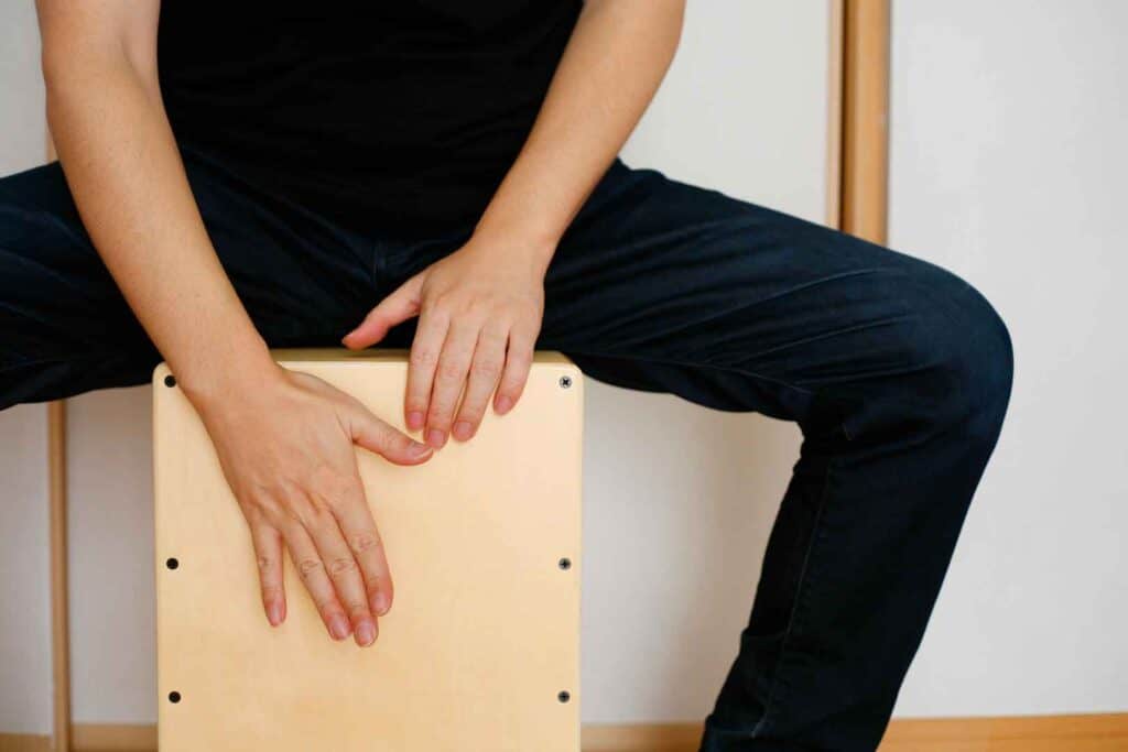 Nahaufnahme der Hände einer Person, die auf einer Cajón (Kistentrommel) sitzt und spielt, mit schwarzer Kleidung und einem hellen Hintergrund.