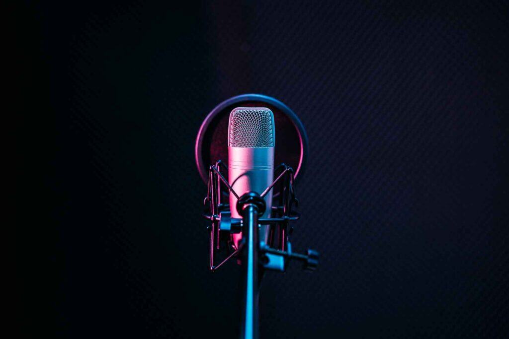 Ein Kondensatormikrofon auf einem Ständer mit einem Popfilter, beleuchtet von rosa und blauem Licht, vor einem dunklen, schallabsorbierenden Hintergrund.