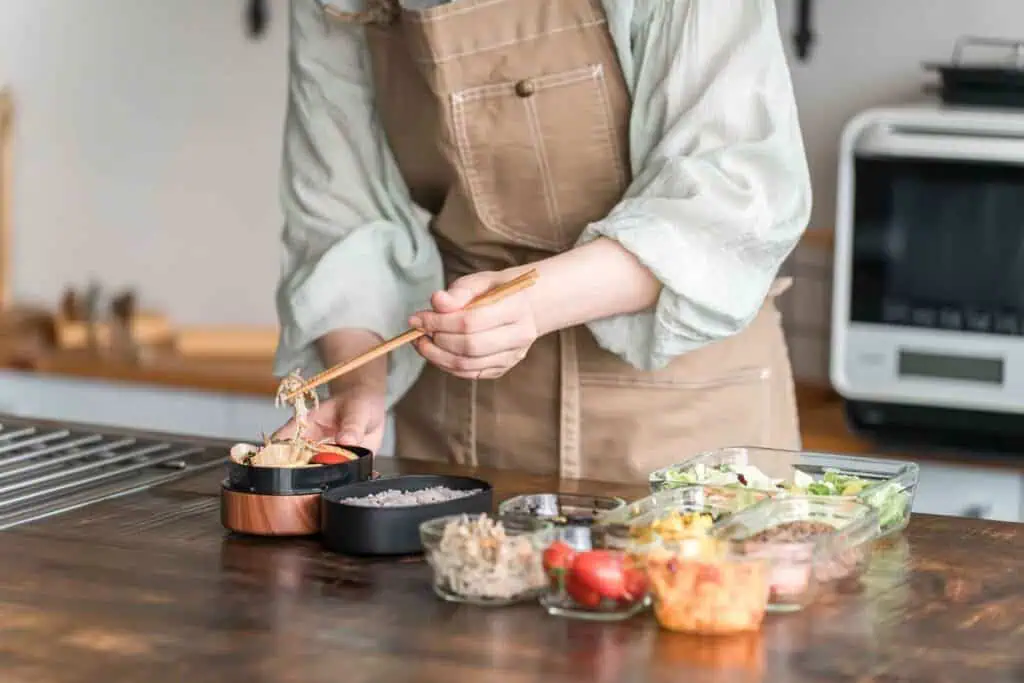 Eine Person in einer Schürze bereitet Mahlzeiten zu, indem sie mit Essstäbchen Essen in eine Bento-Box füllt, während mehrere Glasschalen mit vorbereiteten Lebensmitteln auf einer Holztheke stehen.