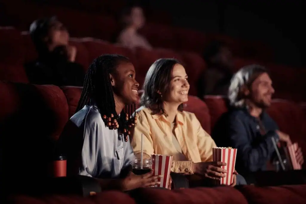 Zwei lächelnde Frauen sitzen in einem Kinosaal und schauen nach rechts, wobei die eine einen Becher und die andere eine Popcorntüte hält.