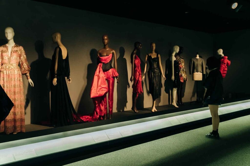 Eine Reihe von Mannequins, die verschiedene Kleider ausstellen, darunter ein langes schwarzes Kleid und ein rotes Korsettkleid, mit einer Person, die die Ausstellung auf der rechten Seite betrachtet.