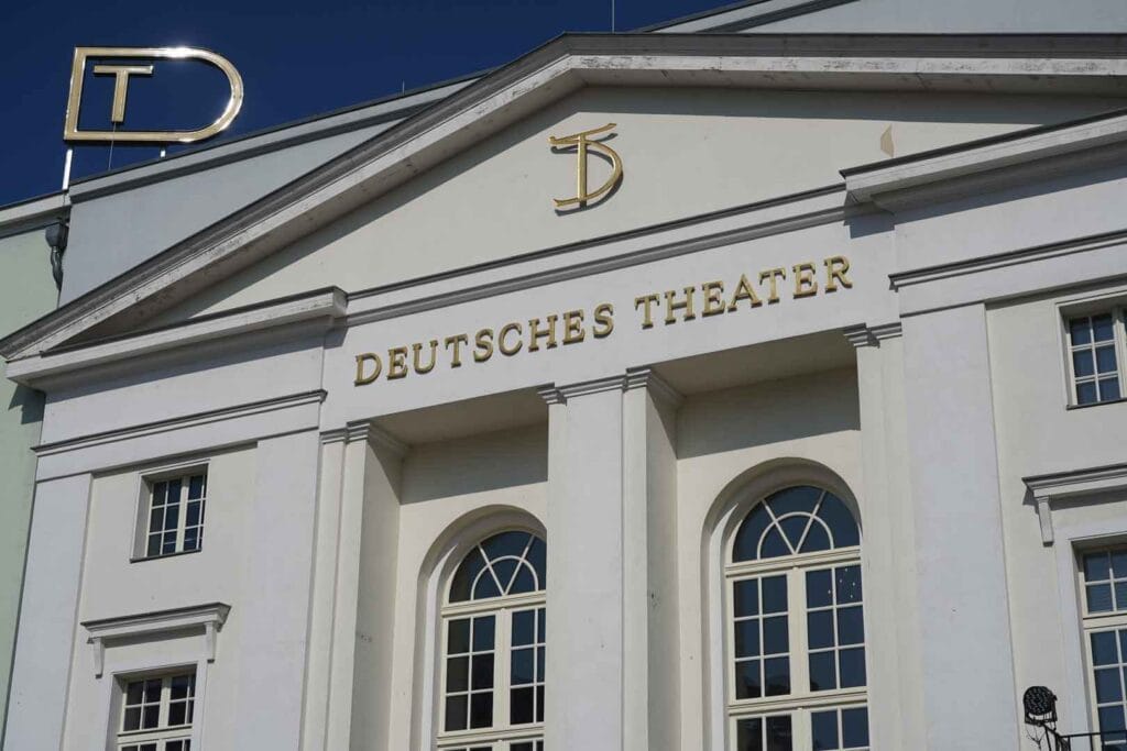 Die Fassade des Deutschen Theaters in Berlin, ein weißes Gebäude mit Säulen und der goldenen Aufschrift "DEUTSCHES THEATER", vor blauem Himmel. Oben auf dem Gebäude sind große goldene Initialen "DT" zu sehen.