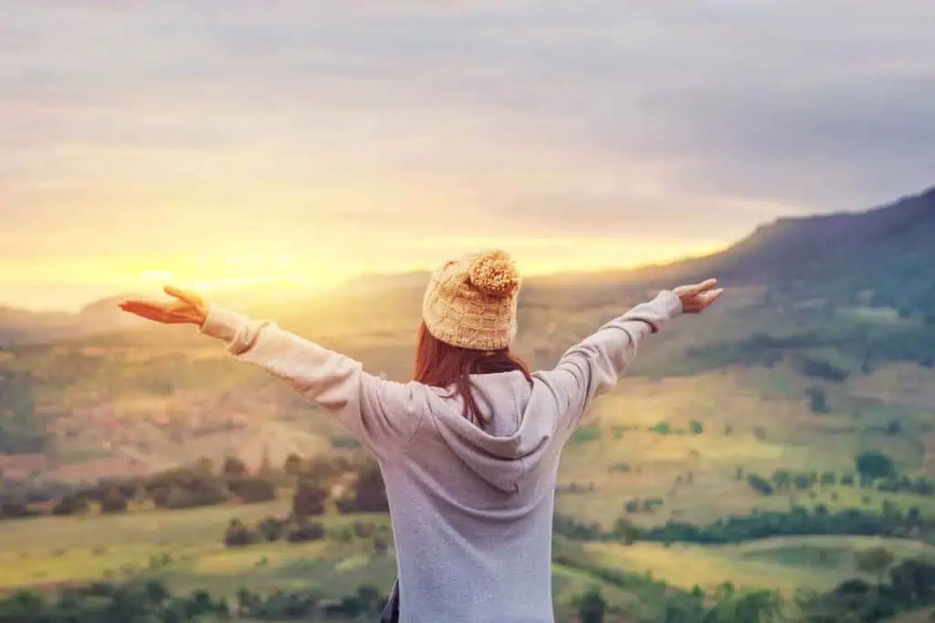 Rückenansicht einer Person mit einer Pudelmütze und einer Kapuzenjacke, die die Arme ausgebreitet hat und auf eine bergige Landschaft bei Sonnenuntergang oder Sonnenaufgang blickt.