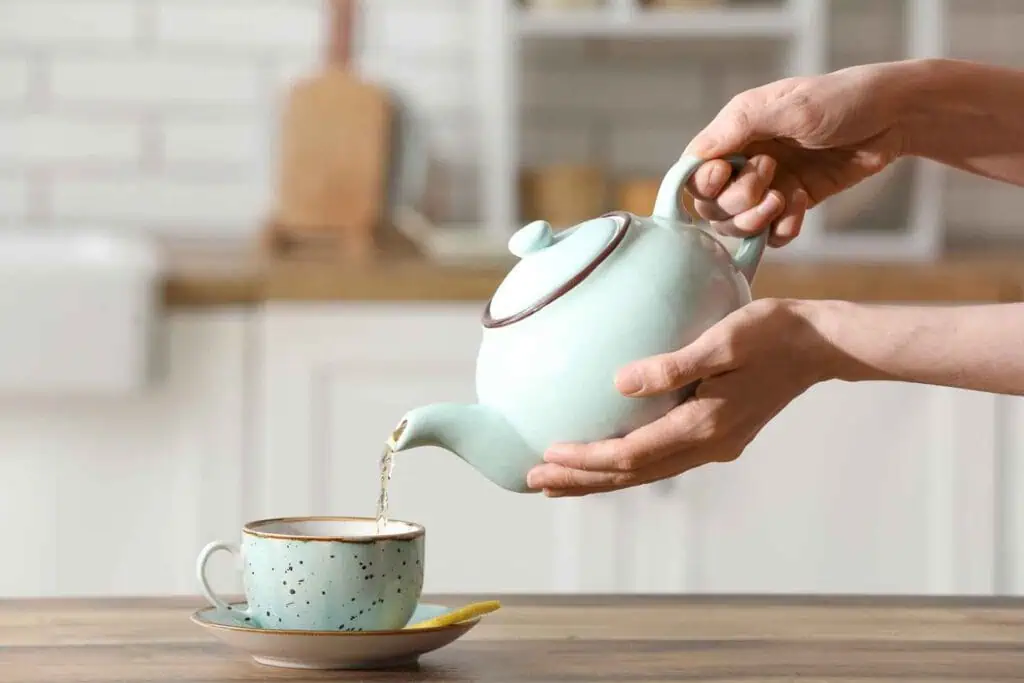 Hände, die eine hellblaue Teekanne halten und Tee in eine passende Tasse gießen, die auf einer Untertasse auf einem Holztisch steht, mit einer unscharfen Küchenzeile im Hintergrund.
