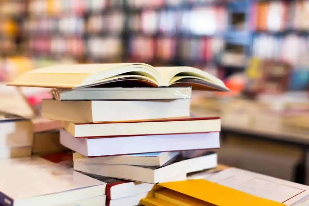 Ein Stapel Bücher auf einem Tisch, mit einem aufgeschlagenen Buch obenauf, vor einem unscharfen Hintergrund von Bücherregalen.