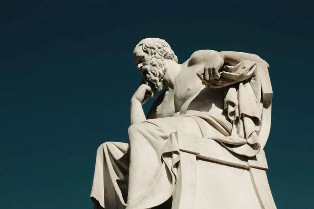 Eine Marmorstatue eines bärtigen Mannes in nachdenklicher Pose, sitzend, mit der Hand am Kinn, vor einem tiefblauen Himmel.