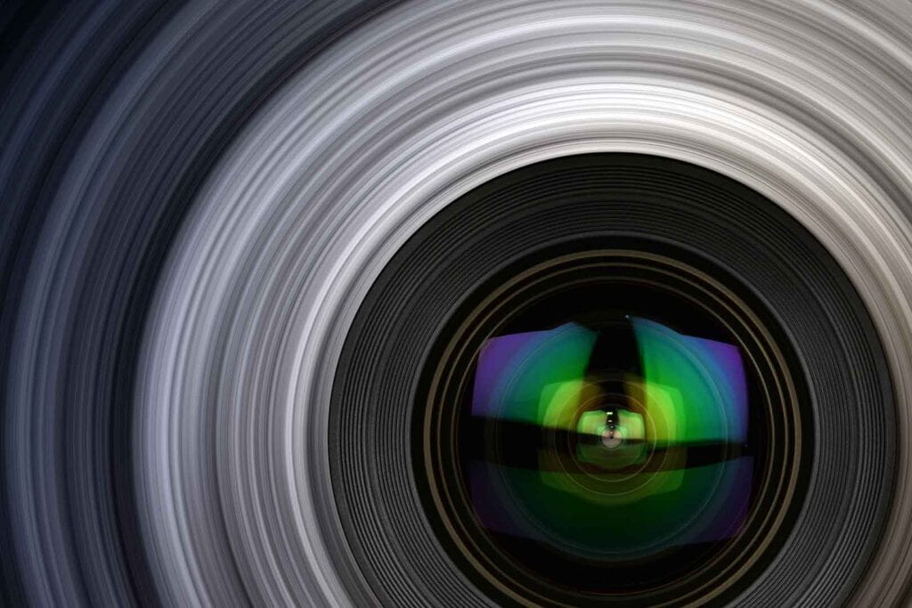 Nahaufnahme des Objektivs einer Kamera mit farbigen Reflexionen auf der Linse und einem spiralförmigen Bewegungsunschärfe-Effekt im Hintergrund.