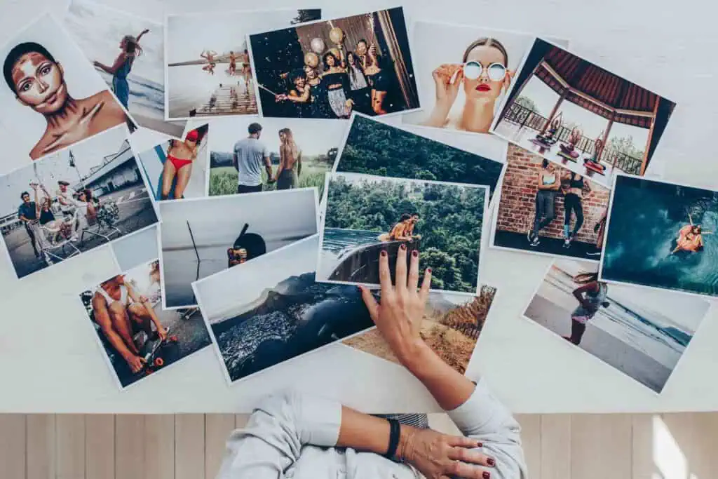 Eine Person legt die Hand auf eine Ansammlung von ausgedruckten Fotos, die auf einer hellen Oberfläche verstreut sind und verschiedene Szenen und Porträts zeigen.