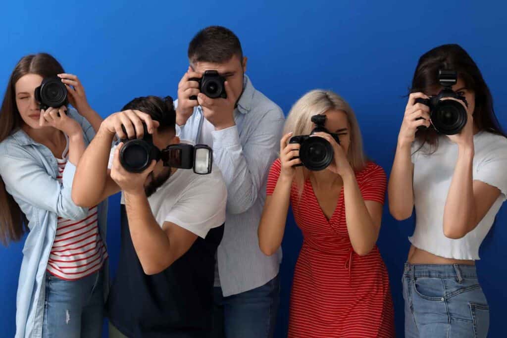 Fünf junge Leute halten Kameras vor ihren Gesichtern und scheinen Fotos zu machen, vor einem blauen Hintergrund.